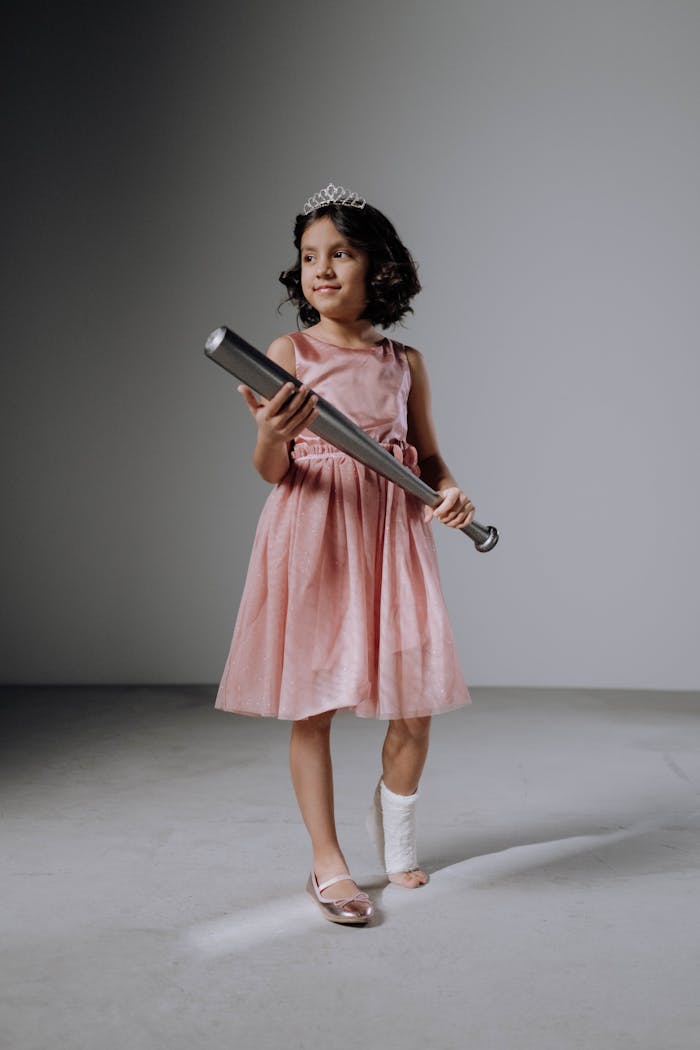 A young girl with a leg cast holding a baseball bat, wearing a pink dress and tiara, standing confidently.